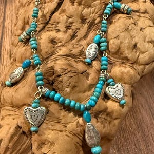 May include: A silver and turquoise necklace with a delicate chain. The necklace features small turquoise beads, silver heart-shaped pendants, and other silver charms. The necklace is displayed on a piece of wood.