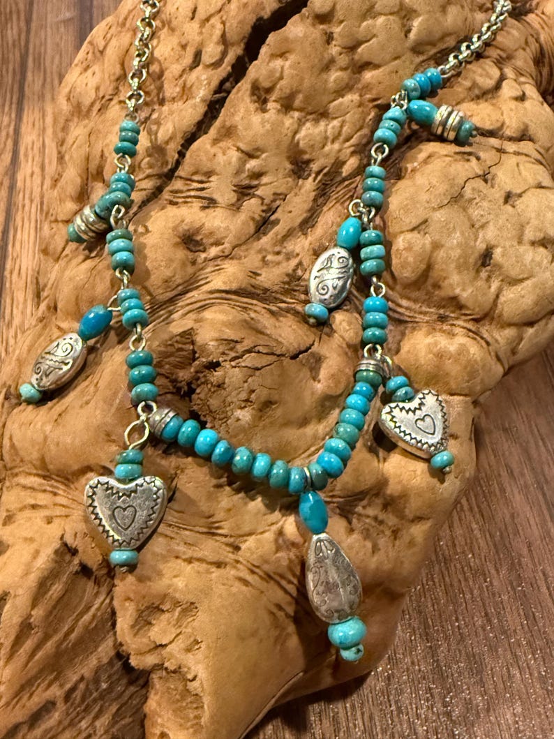 May include: A silver and turquoise necklace with a delicate chain. The necklace features small turquoise beads, silver heart-shaped pendants, and other silver charms. The necklace is displayed on a piece of wood.