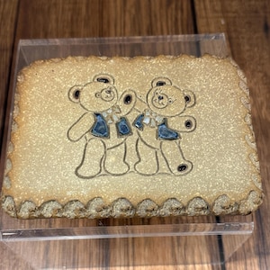 May include: A rectangular ceramic plaque with a light tan background and a scalloped edge. Two cartoon teddy bears with blue vests and scarves are featured on the plaque. The bears are embracing each other.