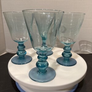 May include: Four teal-colored stemmed wine glasses. The glasses have a flared bowl design and sit atop a round base. The stems feature a series of rounded shapes. The glasses are arranged on a white and blue cake stand.