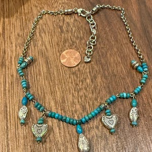 May include: Silver-toned necklace with turquoise beads and heart-shaped charms. The necklace features a chain with a lobster clasp and several dangling charms. The charms have a heart design with a small heart in the center. The necklace is displayed on a wooden surface.