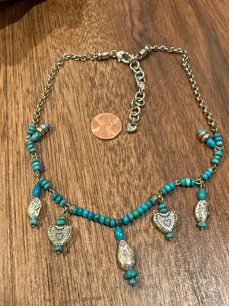 May include: Silver-toned necklace with turquoise beads and heart-shaped charms. The necklace features a chain with a lobster clasp and several dangling charms. The charms have a heart design with a small heart in the center. The necklace is displayed on a wooden surface.