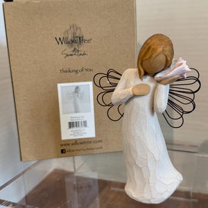 May include: A Willow Tree angel figurine in a white dress, holding a pink and white object. The angel has brown hair and black wire wings. The box behind the angel reads "Thinking of You."