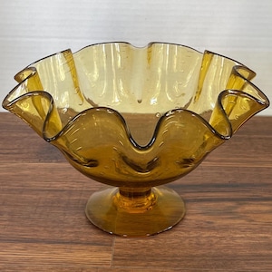 May include: Amber-colored glass bowl with a ruffled rim and pedestal base. The bowl has a wavy, organic shape and is adorned with small air bubbles throughout the glass. The bowl is a decorative piece.