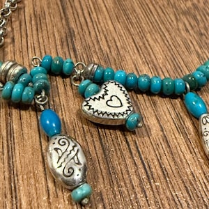 May include: A silver necklace with turquoise beads and decorative pendants. The necklace features a heart-shaped pendant with a heart cutout and an oval pendant with an etched design. The necklace is displayed on a wooden surface.