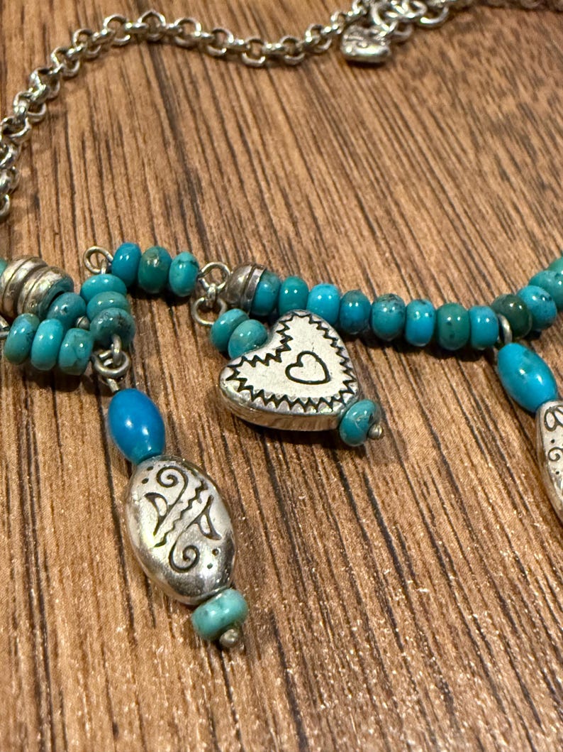 May include: A silver necklace with turquoise beads and decorative pendants. The necklace features a heart-shaped pendant with a heart cutout and an oval pendant with an etched design. The necklace is displayed on a wooden surface.