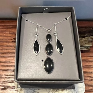 May include: A silver necklace and earrings set with three black teardrop-shaped stones. The jewelry is displayed in a gray box.
