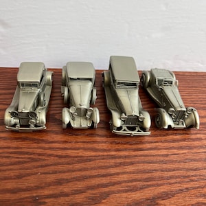 May include: Four miniature pewter model cars in a row on a wooden surface. The cars are all silver in color and appear to be vintage models.