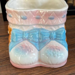 May include: A ceramic planter shaped like a pair of baby booties with a blue bow. The planter is decorated with pink and blue details and has a white glaze.