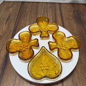 May include: A set of four amber-coloured glass dishes shaped like playing card suits. Three dishes are in the shape of clubs, and one dish is in the shape of a spade. Each glass dish features a textured surface with a central floral pattern.