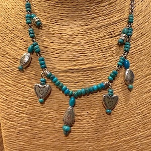 May include: A silver-toned necklace featuring turquoise beads and heart-shaped pendants. The necklace has a chain and several decorative silver elements. The heart pendants have a small heart design in the center.