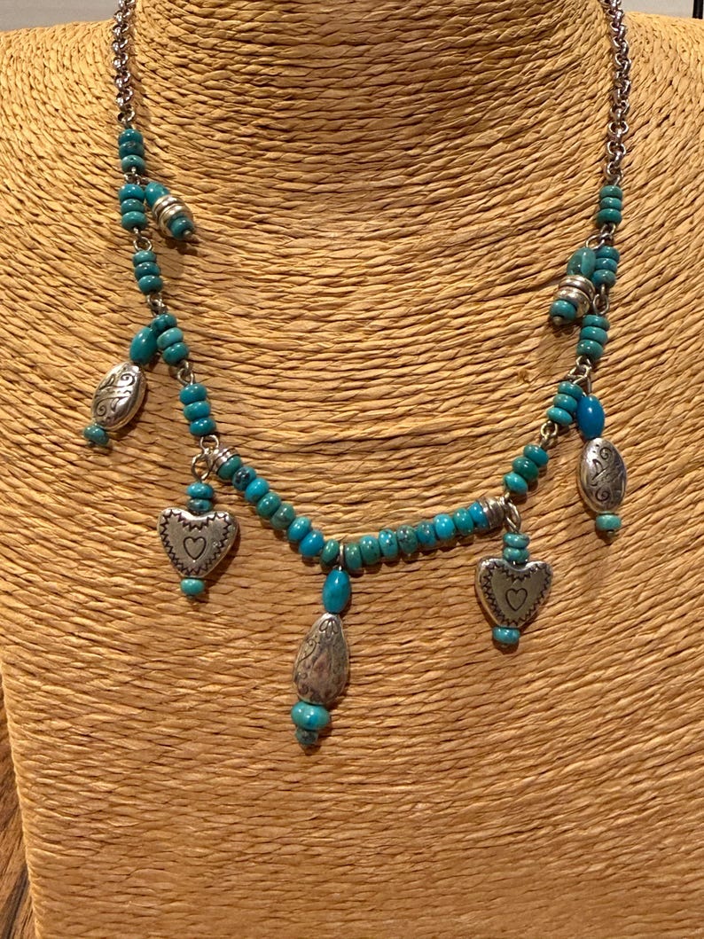 May include: A silver-toned necklace featuring turquoise beads and heart-shaped pendants. The necklace has a chain and several decorative silver elements. The heart pendants have a small heart design in the center.