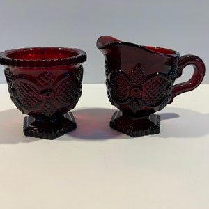 Op de afbeelding: Een robijnrode glazen set van een melkkan en suikerpot. De melkkan heeft een handvat en een tuit, terwijl de suikerpot open is. Beide stukken hebben een decoratief patroon en staan op vierkante voetstukken. De set is waarschijnlijk vintage of antiek.