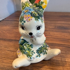 May include: A ceramic bunny figurine with a floral design. The bunny is white with a painted face and is adorned with blue, yellow, and purple flowers and green leaves. The bunny is in a seated position with its paws together.