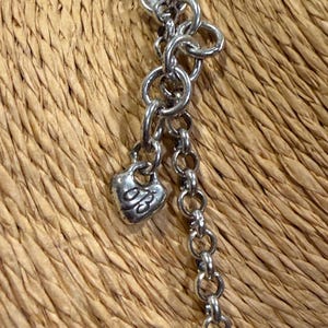 May include: Close-up of a silver-toned necklace with a heart-shaped charm. The charm has an engraved design. The necklace is composed of various chain links and is set against a woven background.