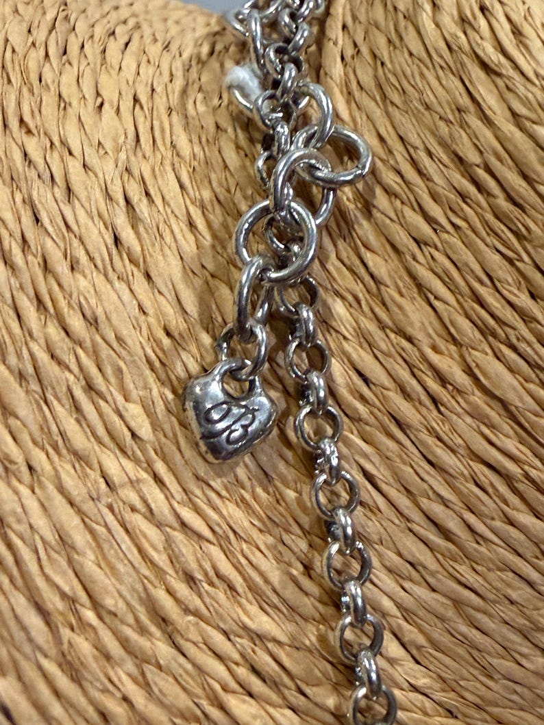 May include: Close-up of a silver-toned necklace with a heart-shaped charm. The charm has an engraved design. The necklace is composed of various chain links and is set against a woven background.