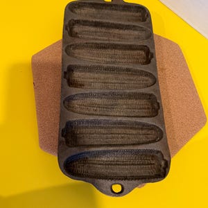 May include: A dark grey cast iron cornbread pan, featuring six corn cob-shaped moulds. The pan is placed on a brown, rectangular surface against a yellow background. It has a handle at each end.