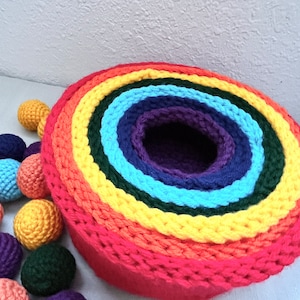 May include: A rainbow-coloured crocheted nesting ring set with a purple centre. The rings are made of yarn in the colours red, orange, yellow, green, blue, and purple. There are also several small crocheted balls in various colours.