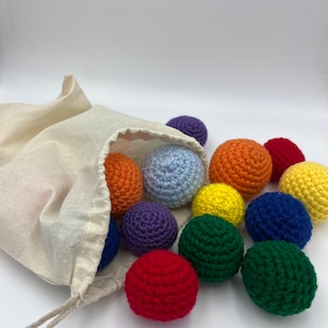 May include: A white drawstring bag filled with colourful crocheted balls. The balls are in a rainbow pattern, with red, orange, yellow, green, blue, purple, and white.