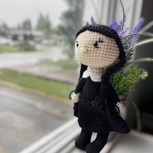 Peut inclure: Une poupée en crochet avec des cheveux noirs, un col blanc et une robe noire. La poupée est debout sur un rebord de fenêtre avec une plante en pot derrière elle.