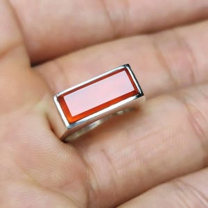 May include: A rectangular gemstone ring with a silver band. The gemstone is a vibrant reddish-orange color, set within a polished silver frame. The ring is shown on a person's finger, highlighting the design and color.