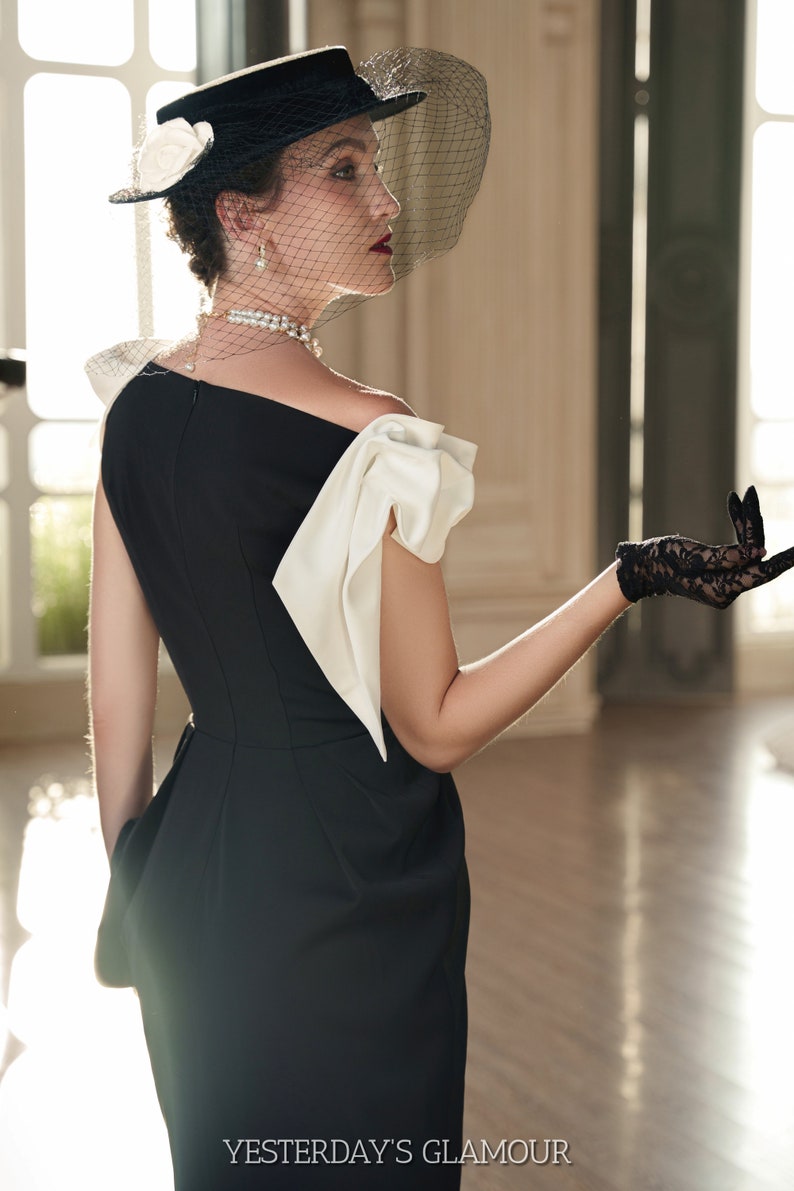 May include: A woman wearing a black dress with a white bow detail on the shoulder, a black hat with a veil, and black lace gloves. The dress has a fitted silhouette and a slightly flared skirt. The woman is standing in a room with a large window in the background. The text "YESTERDAY'S GLAMOUR" is visible at the bottom of the image.