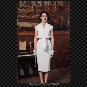 May include: A white sleeveless dress with a bow tie and a white skirt. The dress is fitted and has a cinched waist. The model is wearing white gloves and standing in front of a vintage piano.