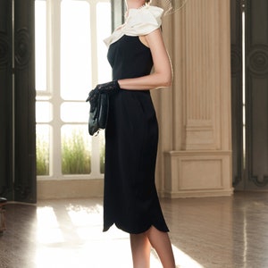 May include: A woman wearing a black dress with a white ruffle collar, black gloves, and a black hat with a veil. She is standing in a room with a large window and a white column behind her. She is wearing beige heels.