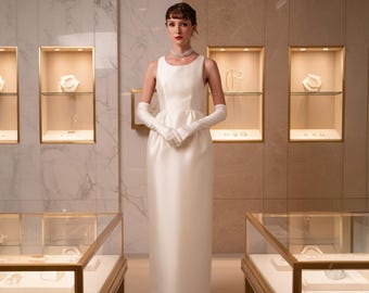 May include: A woman in a white sleeveless gown and long white gloves stands in a jewelry store. She wears a pearl necklace. Display cases with jewelry are in the background. The store has a marble and gold color scheme.