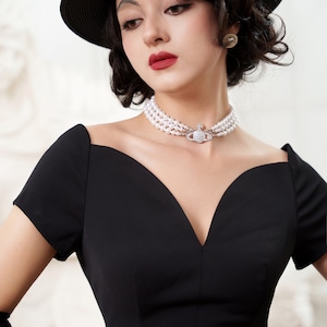 Puede incluir: Una mujer con un vestido negro con un escote pronunciado y un sombrero negro. Lleva un collar de perlas con un colgante de plata. El texto "YESTERDAY'S GLAMOUR" es visible en la parte inferior de la imagen.