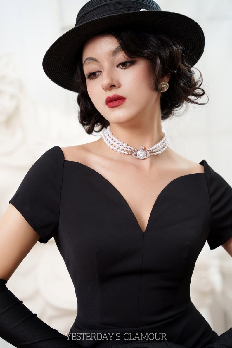 Puede incluir: Una mujer con un vestido negro con un escote pronunciado y un sombrero negro. Lleva un collar de perlas con un colgante de plata. El texto "YESTERDAY'S GLAMOUR" es visible en la parte inferior de la imagen.