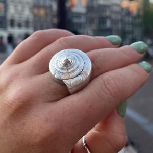 May include: A silver ring with a spiral seashell design. The ring is worn on a hand with green nail polish, with a second, smaller ring on another finger. The background shows a blurred cityscape.