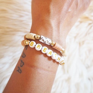Peut inclure: Deux bracelets sur un poignet. Le bracelet du haut est composé de perles brunes et blanches. Le bracelet du bas est composé de perles blanches avec le mot "breathe" écrit en lettres dorées.