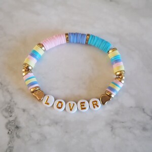 Op de afbeelding: Een kleurrijke kralen armband met een gouden hartbedel aan elk uiteinde. De armband spelt het woord "LOVER" in gouden letters op witte kralen.