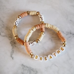 以下が含まれることがあります： ベージュ、白、金色のビーズが付いたブレスレットが2つ。1つのブレスレットには、金色の文字で「BREATHE」と綴られた白いビーズがあります。