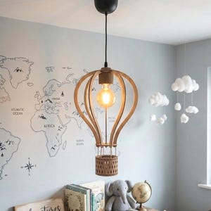 Peut inclure: Une suspension de plafond en forme de montgolfière, fabriquée à partir de lattes de bois clair avec une ampoule centrale de style Edison. Le luminaire est suspendu à un support de plafond noir par un cordon fin, avec une nacelle en bois.