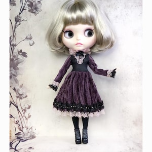 May include: A doll with a silver wig and large eyes, wearing a dark purple velvet dress with black lace trim. The dress has long sleeves with lace cuffs and a black bodice. The doll is also wearing black tights and boots.
