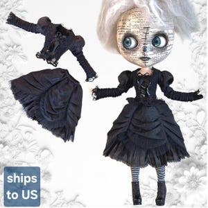 May include: A doll with white hair and a face covered in printed newspaper text, wearing a black dress with puffed sleeves, a ruffled skirt, and black boots. A separate black long-sleeved top and a matching ruffled black skirt are also displayed.