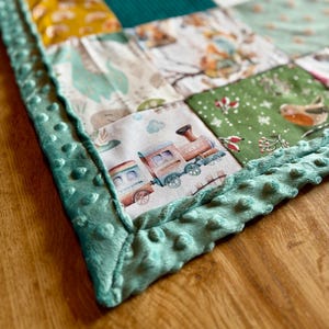Könnte beinhalten: Ein Patchwork-Quilt mit Stoffquadraten in Grün, Gelb, Rosa und Weiß. Der Quilt zeigt eine Zugillustration auf einem Quadrat und einen strukturierten, türkisfarbenen Rand. Er besteht aus verschiedenen Stoffen.