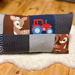 Könnte beinhalten: Rechteckiges Patchwork-Kissen mit einem Reh mit Blume, einem roten Traktor und einem grünen Traktor. Das Kissen hat eine Vielzahl von Stoffquadraten in Grau, Marine, Braun und gepunkteten Mustern. Das Kissen liegt auf einer weißen, flauschigen Oberfläche.