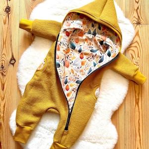 Könnte beinhalten: Senfgelber Baby-Overall mit Kapuze und durchgehendem Reißverschluss. Das Innenfutter zeigt ein farbenfrohes Blumenmuster mit orangefarbenen, blauen und braunen Blättern. Der Overall liegt auf einer weißen, flauschigen Oberfläche.