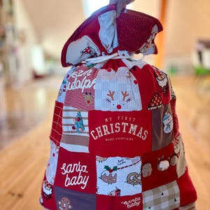 Könnte beinhalten: Ein weihnachtlicher Geschenkesack im Patchwork-Design aus verschiedenen roten, weißen und grauen Stoffquadraten mit Weihnachtsmotiven. Der Sack trägt die Aufschriften My First Christmas, Merry Everything und Santa Baby und hat einen weißen Kordelzug.