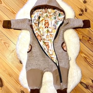 Könnte beinhalten: Beige Baby-Overall mit Kapuze und braunen Akzenten. Das Innenfutter zeigt einen Waldprint mit Bären, Füchsen und anderen Tieren. Der Overall hat einen durchgehenden Reißverschluss und braune Bündchen.