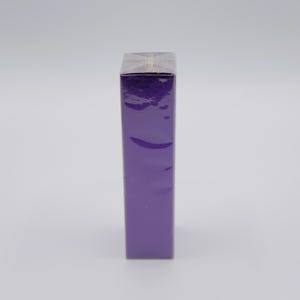 May include: A tall, rectangular, purple box wrapped in clear cellophane. The box is standing upright against a white background. The cellophane is crinkled, and the box is a solid, vibrant purple color.
