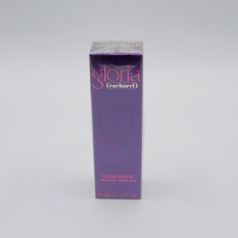 May include: A rectangular perfume box in shades of purple. The box features the name "gloria (cacharel)" in pink script. The bottom of the box reads "eau de toilette" and "e 30 ml - 1.0 fl oz."