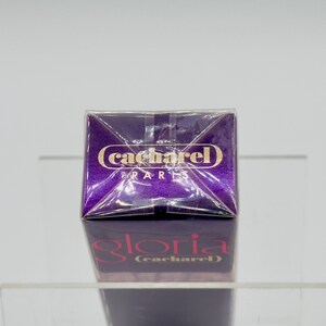 May include: A purple perfume box with the brand name "cacharel PARIS" in gold lettering. The box is reflected on a clear surface, with the name "gloria (cacharel)" visible below. The packaging is sealed.
