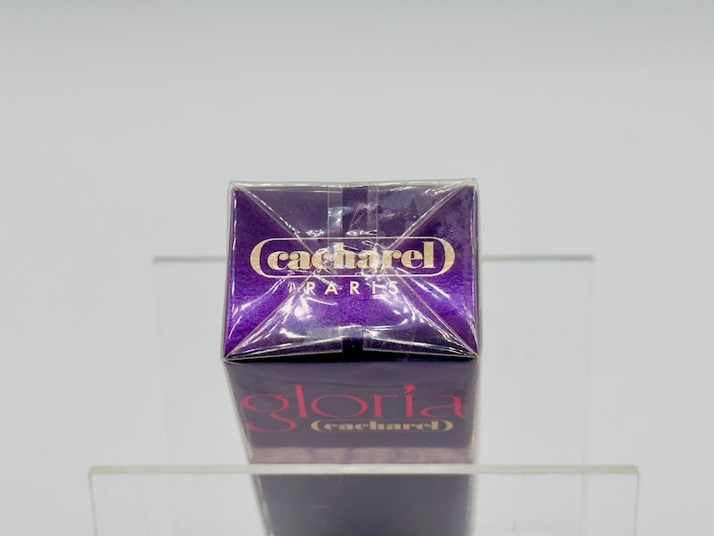 May include: A purple perfume box with the brand name "cacharel PARIS" in gold lettering. The box is reflected on a clear surface, with the name "gloria (cacharel)" visible below. The packaging is sealed.