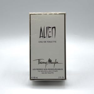 Könnte beinhalten: Eine rechteckige Schachtel Thierry Mugler Alien Eau de Toilette. Die Schachtel ist hellgrau mit schwarzem Text, der "ALIEN" und "EAU DE TOILETTE" lautet. Auf der Unterseite der Schachtel steht "60 ml" und "2 Fl. Oz."