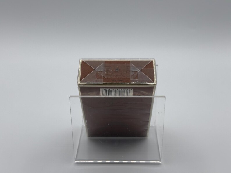 May include: A brown rectangular box with a silver trim, displayed on a clear acrylic stand. The box is sealed with clear tape and has a barcode on the side. The background is a plain, light gray.