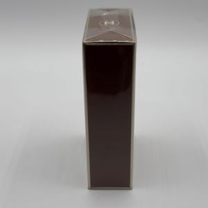 May include: A rectangular, dark brown box with a clear plastic top. The box has a glossy finish and a small, circular logo on the top. The box is standing upright against a white background.